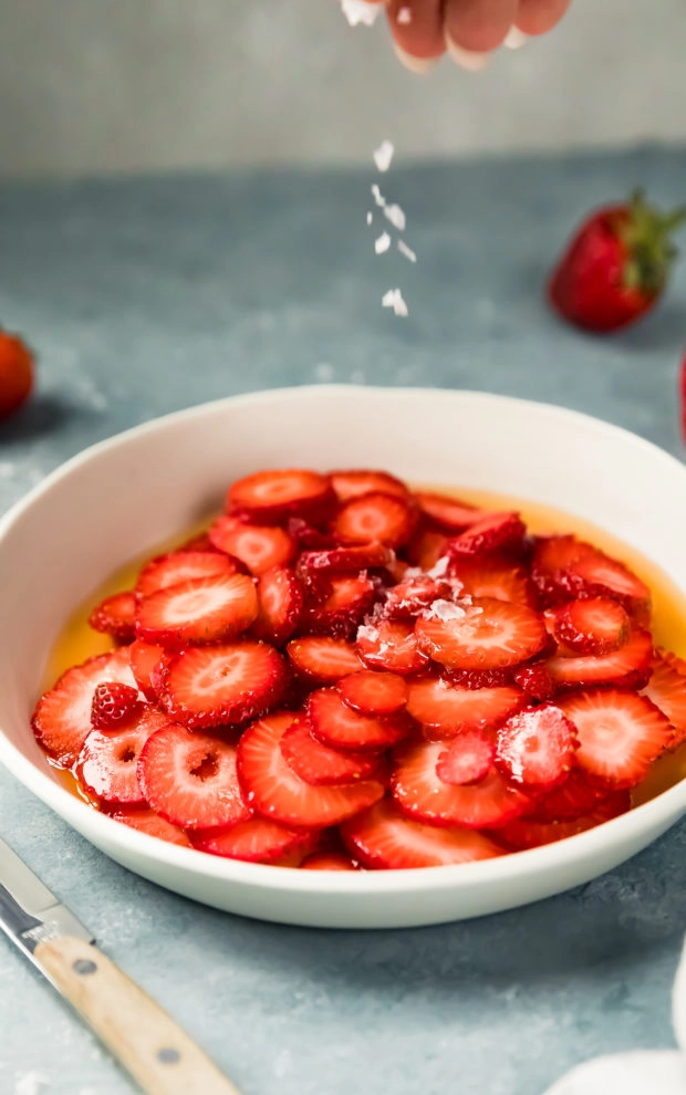Angled shot of sliced strawberries in a white shallow bowl with hand sprinkling coarse salt over the strawberries; with fresh strawberries, a wooden handled knife and neutral linen surrounding the bowl. (photo of the second part of step one of the recipe)