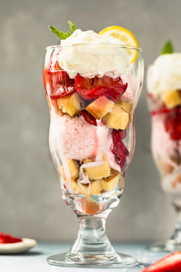 Straight on, up-close shot of a homemade Strawberry Ice cream Sundae with another sundae blurred and slightly visible in the background and fresh strawberries surrounding the sundae glass.
