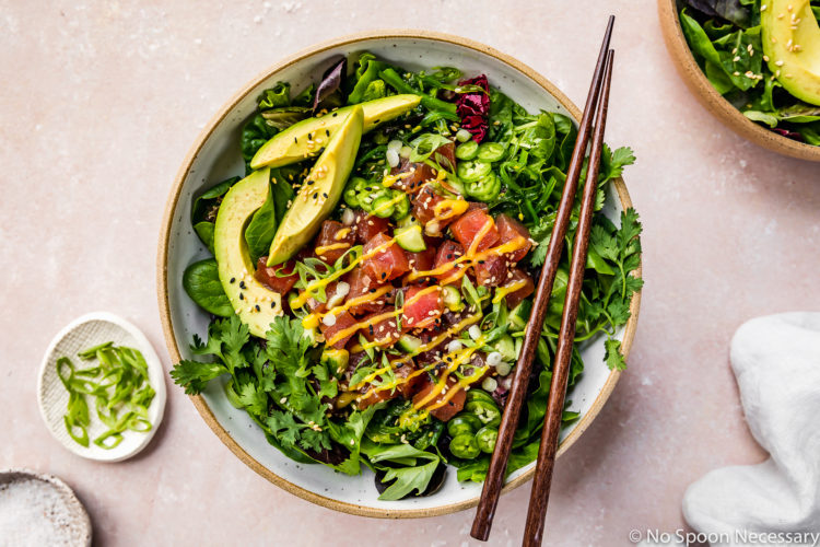 Overhead shot of an Avocado Mango Tuna Poke Salad Bowl with wood chopsticks resting on top of the bowl and an additional bowl and ramekins of sliced scallions and sea salt surrounding the bowl and barely visible in the shot.