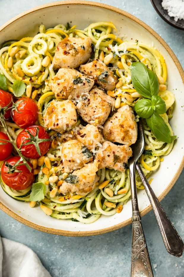 Overhead, close-up photo of Basil Pesto Chicken Pasta garnished with oven roasted tomatoes in a large bowl with two forks tucked under the pasta.
