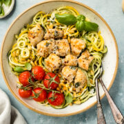 Overhead, landscape photo of Basil Pesto Chicken Pasta in a large bowl with oven roasted tomatoes and two forks inserted into the noodles; with a small jar of pesto, neutral linen, and ramekins of salt and basil surrounding the bowl.