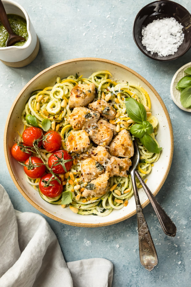 Overhead photo of Pesto Chicken Pasta in a large bowl with oven roasted tomatoes and two forks inserted into the noodles; with a small jar of pesto, neutral linen, and ramekins of salt and basil surrounding the bowl.