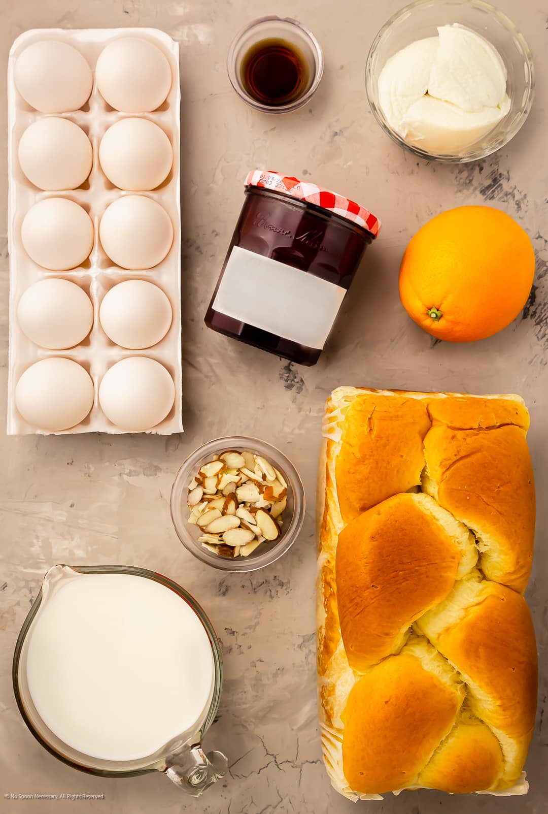A loaf of brioche bread, sweet strawberry jam, eggs, ricotta cheese, vanilla, and heavy cream neatly organized on a kitchen counter.