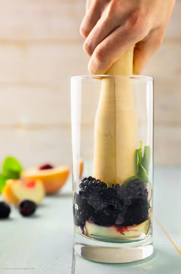 Action photo of a person muddling peaches, mint leaves, and blackberries in a tall glass.