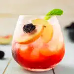 Straight on photo of an elderflower cocktail garnished with peach slices, blackberries, and mint in a short glass.