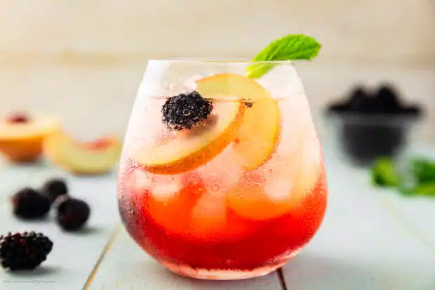 Straight on photo of an elderflower cocktail with vodka, peach slices, blackberries, and mint in a short glass.