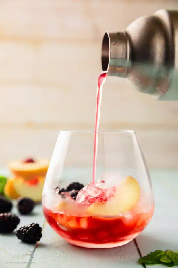Action photo of a peachy cocktail being poured from a shaker into a rocks glass.