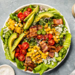 Overhead shot of a Summer Corn BLAT Salad in a white shallow bowl with a small jar of avocado jalapeno dressing, vine ripe cherry tomatoes, and ramekin of salt surrounding the bowl.