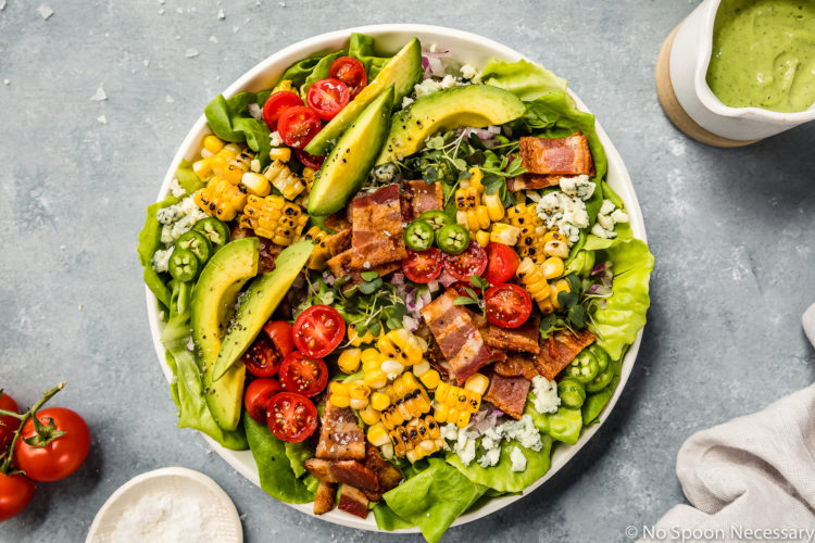 Overhead shot of a Summer Corn BLAT Salad in a white shallow bowl with a small jar of avocado jalapeno dressing, vine ripe cherry tomatoes, and ramekin of salt surrounding the bowl.