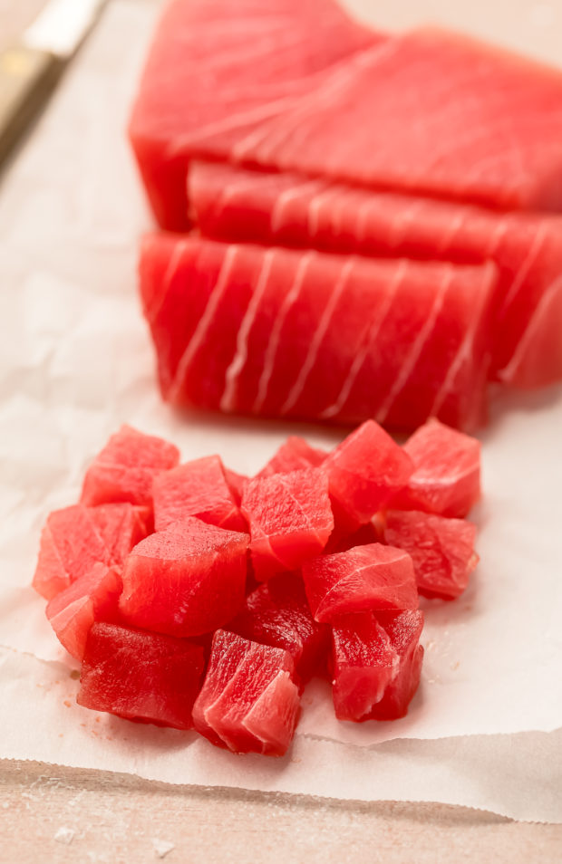 Angled photo of a raw sushi-grade tuna filet partially cut into cubes on a sheet of parchment paper (the first step in the recipe for poke salad).