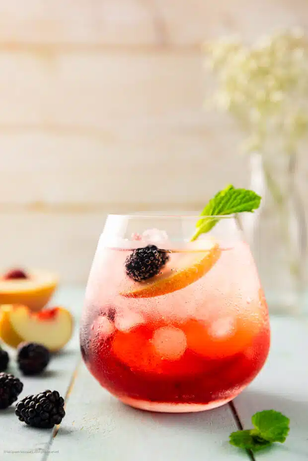 Straight on photo of an elderflower cocktail with peach, vodka, and blackberries in a short glass.