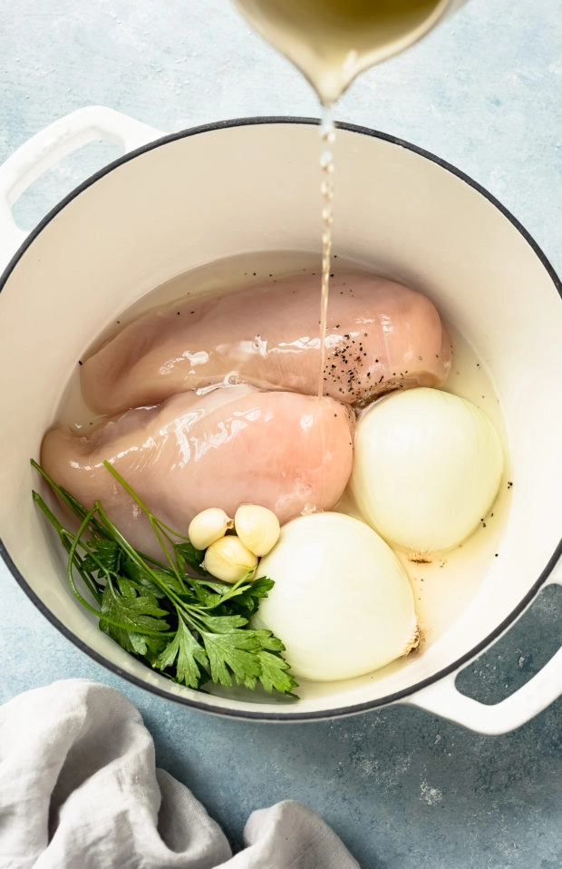 Angled photo of chicken broth being poured into a large white pot containing chicken breasts, garlic cloves, halved white onions and fresh parsley - photo of how to poach chicken for chicken salad.