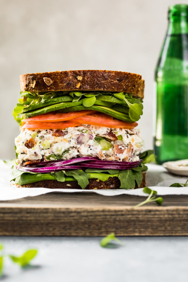 Straight on shot of a Creamy Bacon Everything Spice Chicken Salad Sandwich with lettuce, tomato, avocado and red onions on multigrain bread on a gray wood board with a green bottle of water blurred in the background. 