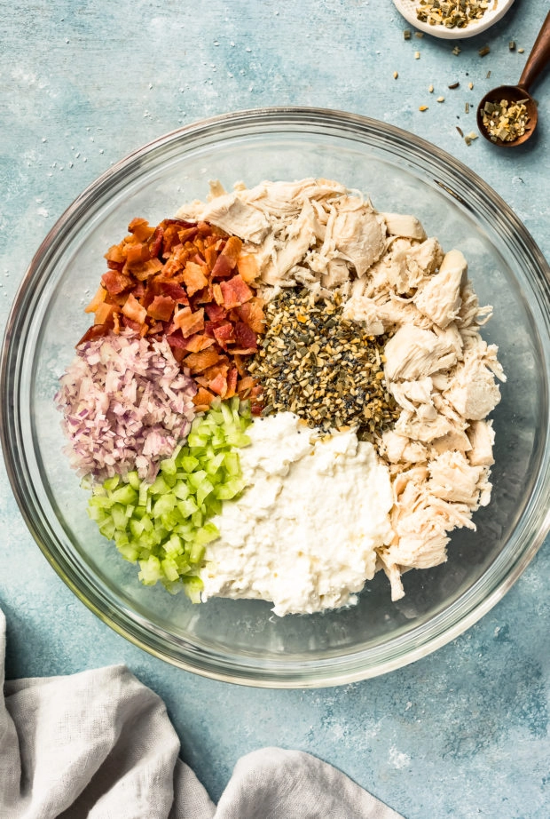 Overhead shot of all the ingredients needed to make Creamy Bacon Everything Spice Chicken Salad neatly organized in a glass bowl with a neutral blue linen and ramekin of everything spice surrounding the bowl. (Photo of step 3 in the Creamy Bacon Everything Spice Chicken Salad recipe.)