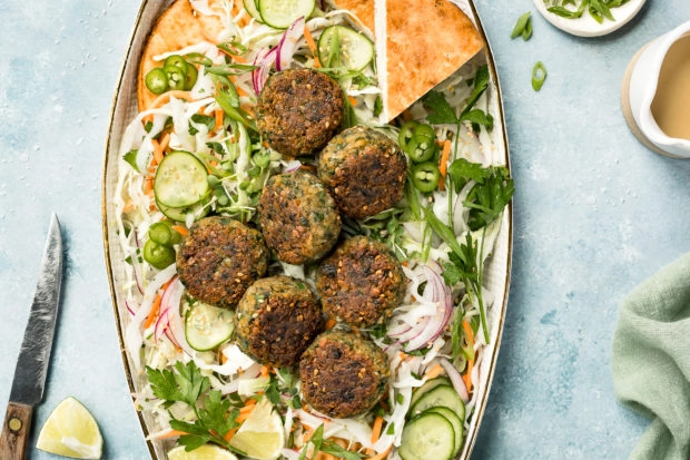 Overhead photo of a platter of Asian Crispy Pan Fried Falafels on a bed of Asian slaw with pita bread; with a pale green linen, knife, lime wedge, ramekin of sliced scallions and small ceramic jar of miso tahini surrounding the platter.