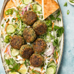 Overhead shot of a platter of Asian Crispy Pan Fried Falafels on a bed of Asian slaw with pita bread; with a pale green linen, knife, lime wedge, ramekin of sliced scallions and small ceramic jar of miso tahini surrounding the platter.