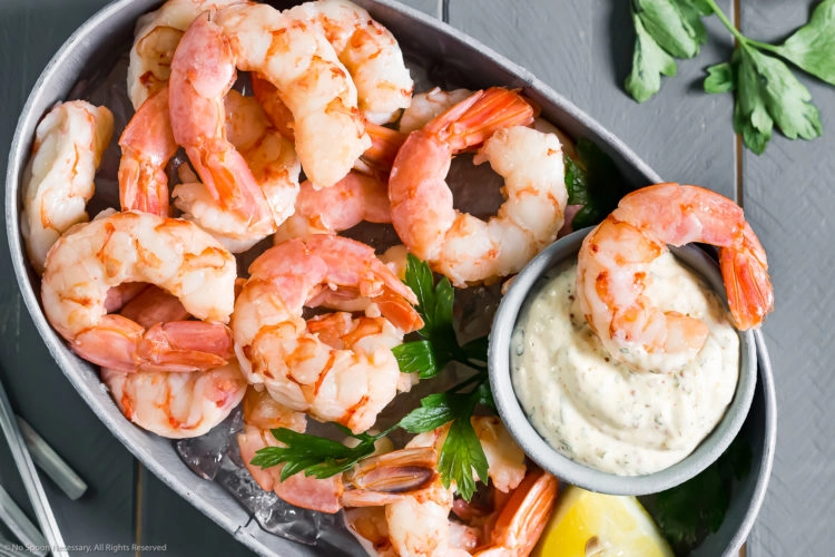 Overhead photo of a small, deep-sided serving bucket filled with ice and topped with Perfect Poached Cocktail Shrimp with a small container of Remoulade sauce next to the shrimp and an individual shrimp dunked into the sauce.