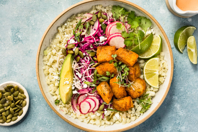 Overhead photo of White Fish Taco Bowls with cauliflower rice and slaw in a white bowl with a ramekin of pepitas, lime wedges and a ramekin of sauce surrounding the bowl.