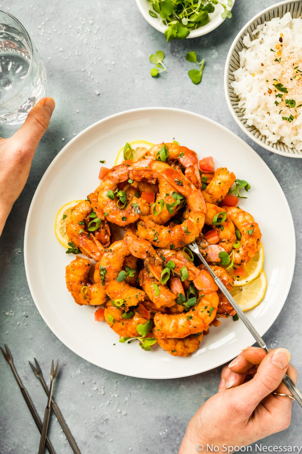 Overhead shot of Easy Cajun Honey Butter Shrimp garnished with slices of lemon and fresh green onions on a white plate with a hand inserting a seafood fork into one of the shrimp and another hand holding a glass of water next to the plate; with a ramekin of microgreens and bowl of white rice arranged around the plate.