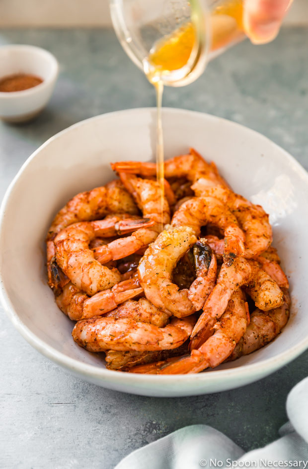 Angled shot of honey marinade being poured over cajun seasoned shrimp in large bowl with a neutral colored linen and a ramekin of cajun seasoning arranged around the bowl - photo of step 2 of the Easy Cajun Honey Butter Shrimp recipe.