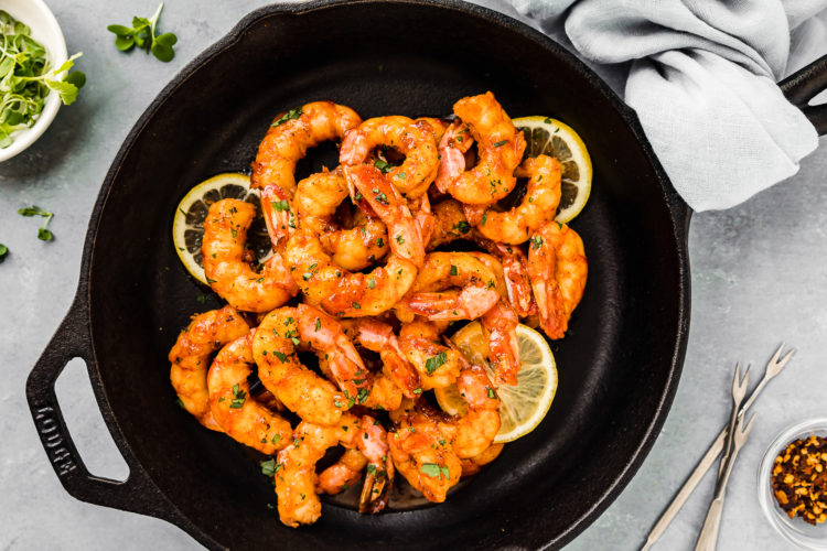 Overhead shot of Easy Cajun Honey Butter Shrimp garnished with slices of lemon in a small cast iron skillet with seafood forks, a neutral colored linen and ramekin of microgreens arranged around the skillet.