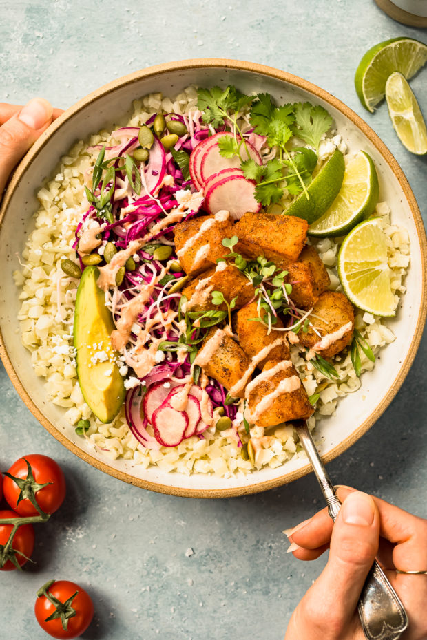 Overhead photo of a fish taco bowl with bite-size pieces of white fish and cauliflower rice in a white bowl with two hands holding the bowl.