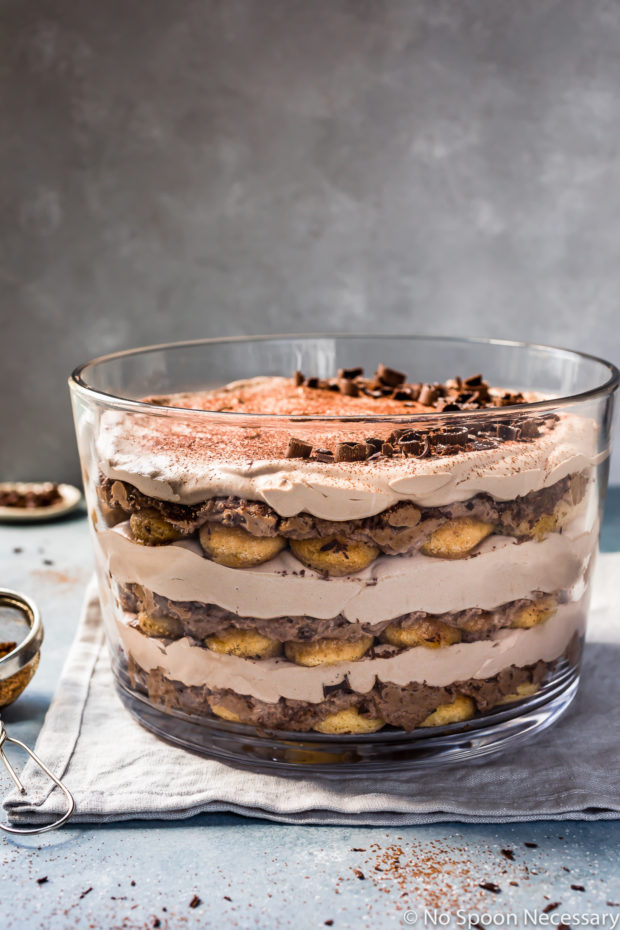 Slightly angled, straight on shot of an Easy Chocolate Tiramisu Trifle on a pale blue linen with a mini fine mesh sifter full of cocoa powder and a small ramekin of shaved chocolate off to the side of the trifle.