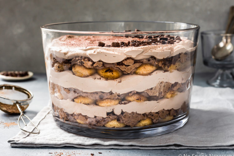 Slightly angled, straight on shot of an Easy Chocolate Tiramisu Trifle on a pale blue linen with a mini fine mesh sifter full of cocoa powder off to the side of the trifle and a small ramekin of shaved chocolate and small glass containing spoons blurred behind the trifle.