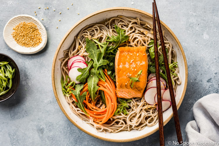 Overhead shot of Green Tea Poached Salmon Soba Noodles with arugula, sliced radishes and julienned carrots in a neutral colored bowl with chopsticks resting on the edge; and a ramekin of sesame seeds, sliced scallions and a pale blue linen arranged around the bowl.