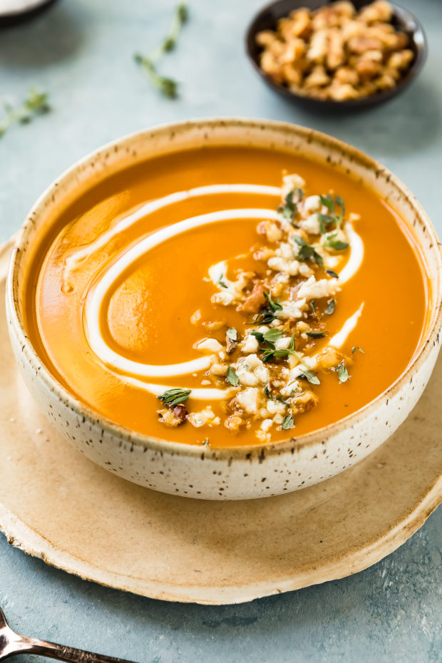 45 degree angle photo Roasted Sweet Potato Soup garnished with walnuts, thyme, gorgonzola and swirls of cream in a neutral colored bowl with a spoon in front of the bowl and sprigs of fresh thyme and a ramekin of walnuts blurred in the background.