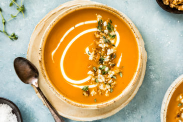 Overhead photo of Creamy Sweet Potato Soup garnished with walnuts, thyme, gorgonzola and swirls of cream in a neutral colored bowl on a plate with sprigs of fresh thyme, a spoon, additional bowl of soup and small brown ramekins of walnuts and salt surrounding the bowl.