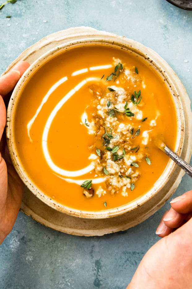 Overhead photo of a hand holding a spoon inserted into creamy sweet potato soup in a neutral colored bowl.