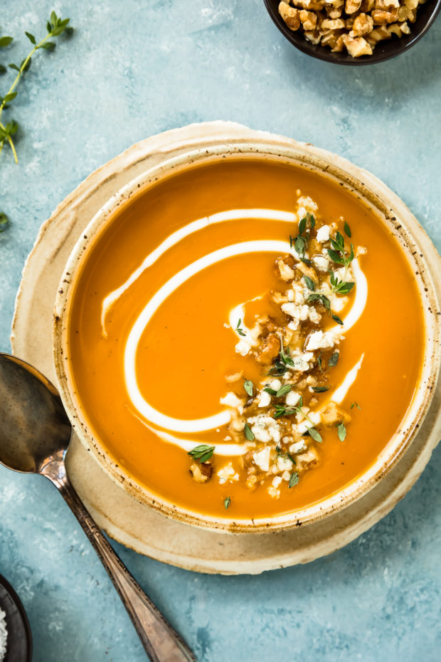 Overhead photo of Creamy Sweet Potato Soup with fresh thyme, walnuts and a cream swirl in a neutral colored bowl with sprigs of fresh thyme, a spoon and small brown ramekins of walnuts and salt surrounding the bowl.
