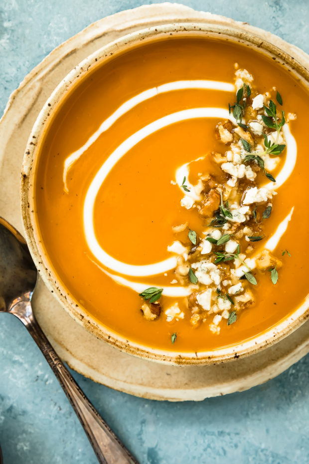 Overhead photo of Sweet Potato Soup garnished with fresh thyme, walnuts and a cream swirl in a neutral colored bowl with a spoon resting next to the bowl.