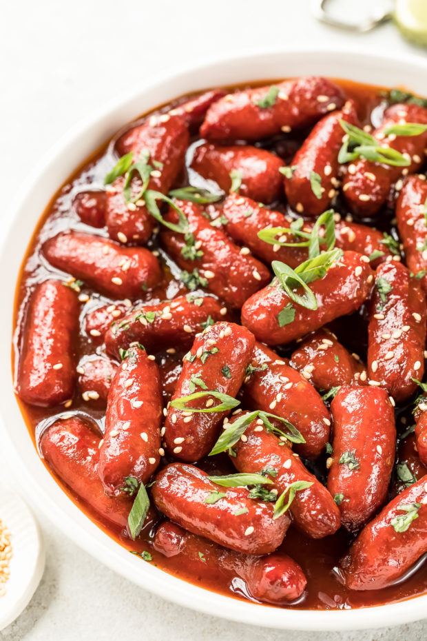Angled shot of a white bowl filled with Slow Cooker Honey Sriracha Little Smokies with a ramekin of sesame seeds, a bottle opener and beer cap blurred in the corners of the shot.