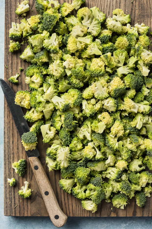 Overhead photo of a head of fresh broccoli cut into bite-size florets on a large wood cutting board with a knife next to the florets.
