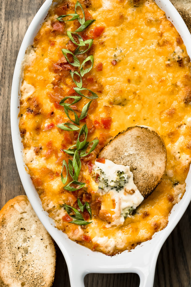 Overhead photo of Baked Broccoli Dip in a white oval baking dish with a slice of toasted baguette inserted into the dip showcasing the creamy interior.