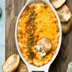 Overhead photo of Broccoli Dip in a white oval baking dish with a slice of toasted baguette inserted into the dip and slices of baguette, a green linen, and ramekins of sliced scallions and salt surrounding the baking dish.