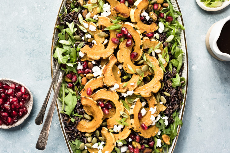 Overhead photo of Wild Rice Salad featuring roasted delicata squash, pomegranate arils and sliced scallions on a white platter with serving spoons inserted into the salad; and ramekins of pomegranate arils, sliced scallions and a small container of honey balsamic vinaigrette arranged around the salad.