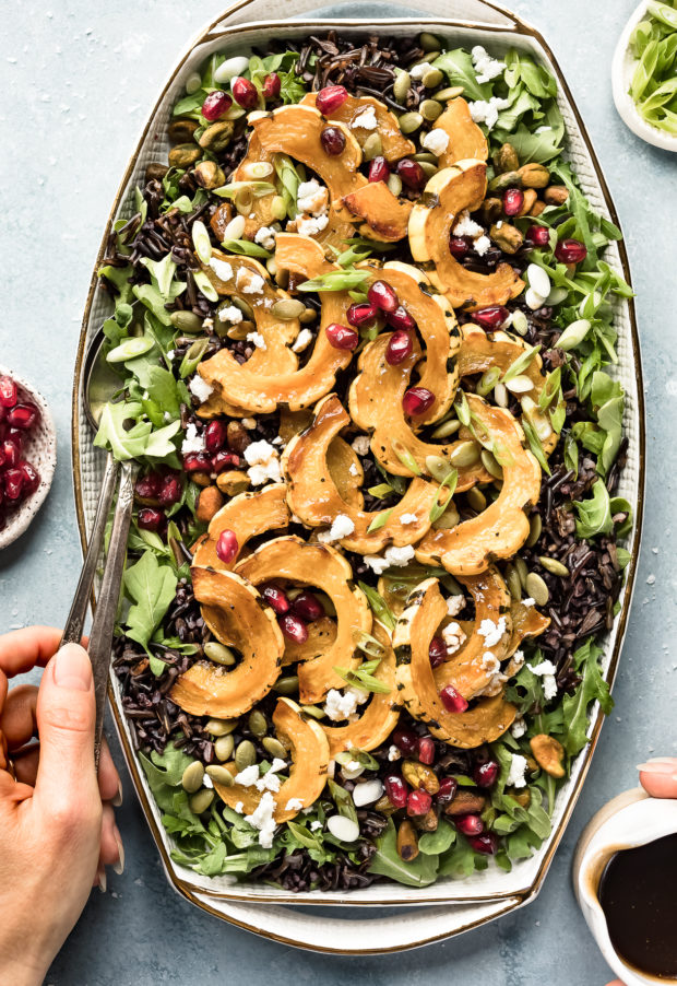 Overhead photo of Wild Rice Salad on a white platter with a hand holding serving spoons inserted into the salad and another hand holding a small container of honey balsamic vinaigrette next to the platter.