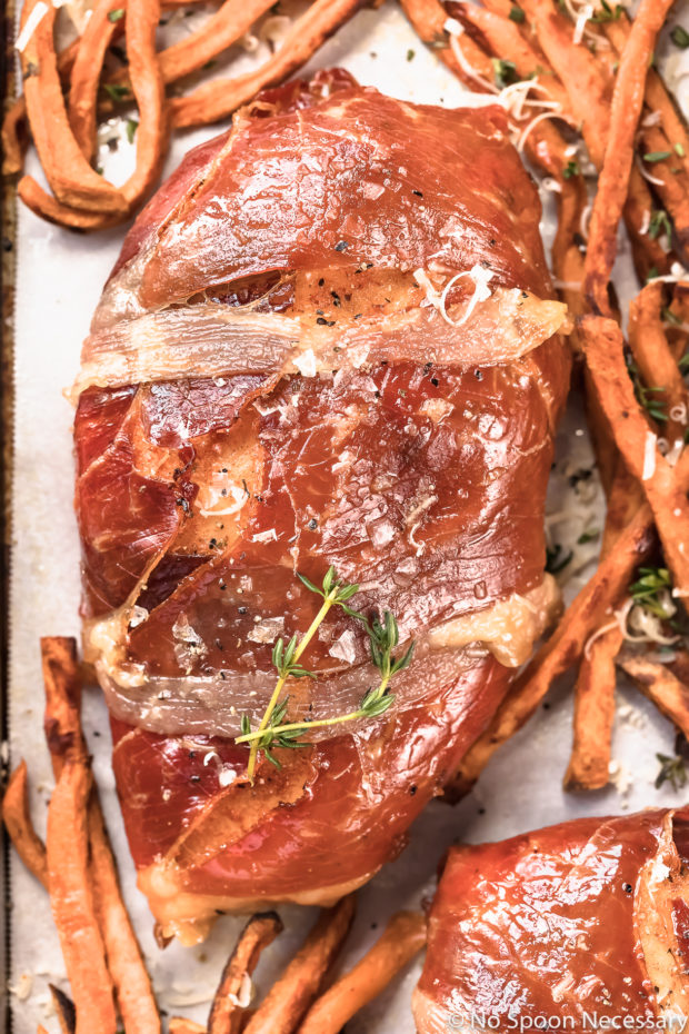 Overhead, up close shot of a Brown Sugar Prosciutto Wrapped Chicken Breast garnished with flaky salt and fresh thyme and surrounded by Sweet Potato Fries on a parchment paper lined sheet pan. 