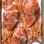 Overhead shot of Brown Sugar Prosciutto Wrapped Chicken & Sweet Potato Fries on a sheet pan with a ramekin of flaky salt; with a light green linen, ramekin of microgreens and two glasses of white wine arranged around the pan.
