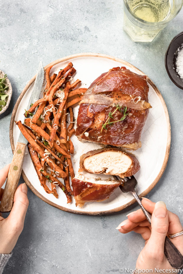 Overhead shot of partially sliced Brown Sugar Prosciutto Wrapped Chicken and Sweet Potato Fries on a white plate with a hand holding a fork tucked under a slice of chicken and another hand holding a steak knife off the other side of the plate; with a glass of white wine and ramekins of salt and microgreens arranged around the plate. 