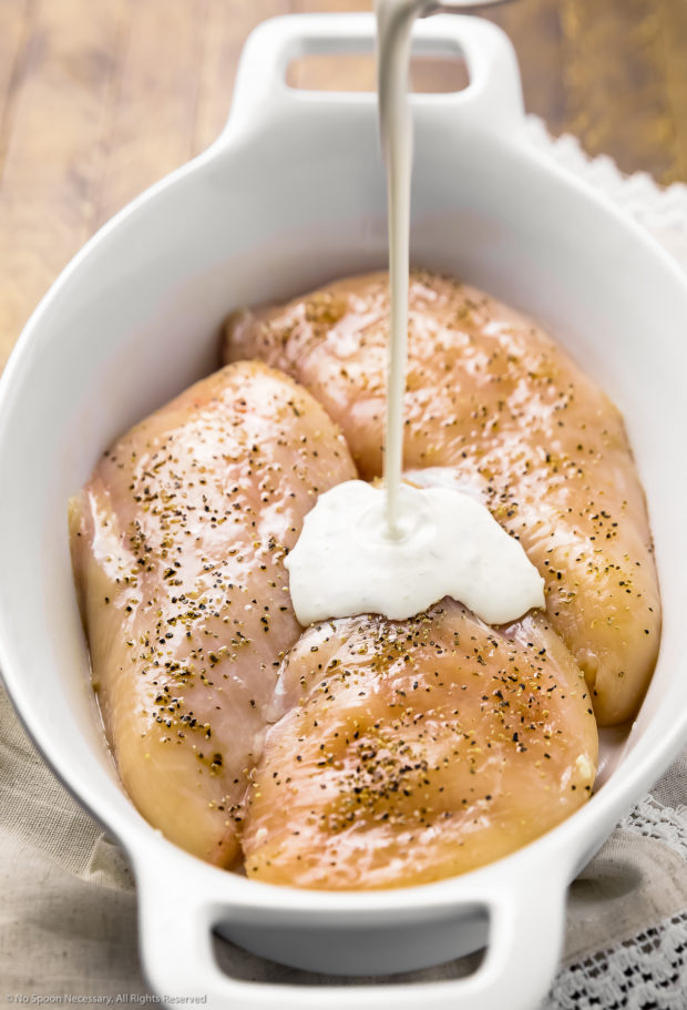 Angled photo of three chicken breasts in a white oval baking dish with heavy cream being poured on top.