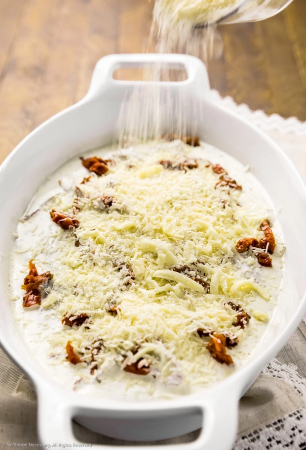 Angled photo of grated parmesan being sprinkled over three raw chicken breasts in a heavy cream and sun-dried tomatoes sauce.