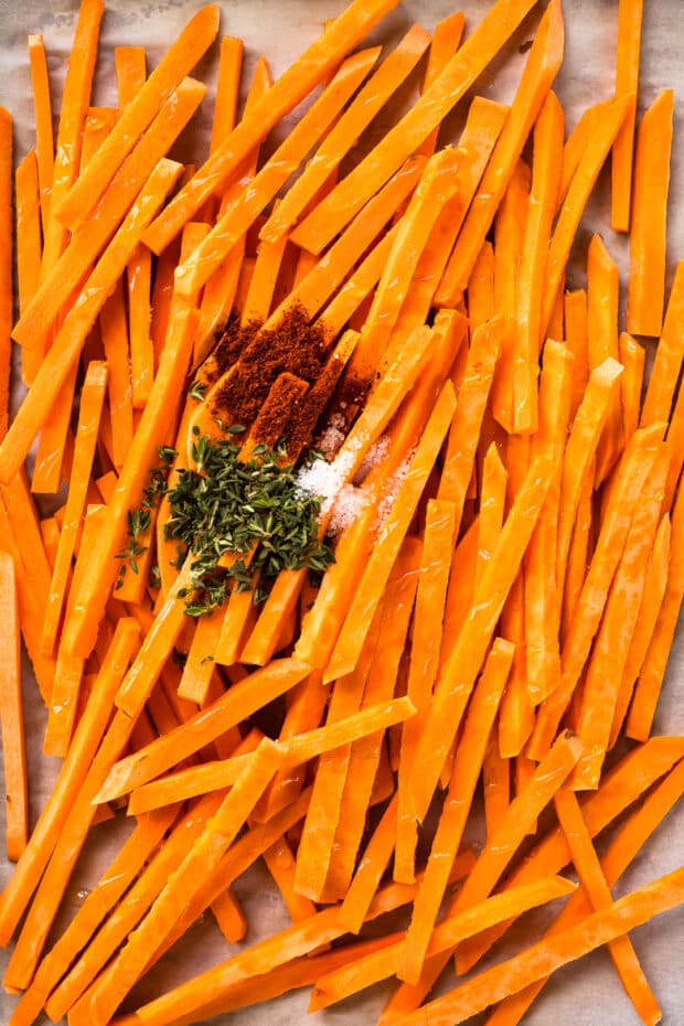 Overhead photo of raw sweet potato fries with salt, paprika, and herbs on a parchment lined sheet pan - before baking.