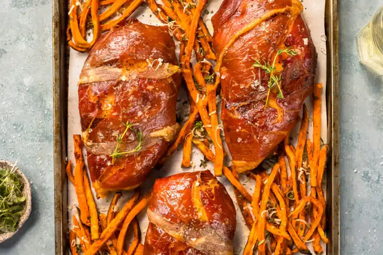 Overhead photo chicken wrapped in prosciutto with crisp sweet potato fries on a baking sheet.
