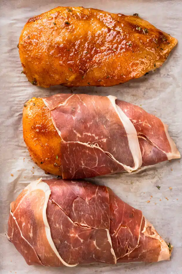 Action photo of prosciutto being wrapped around seasoned chicken breasts.