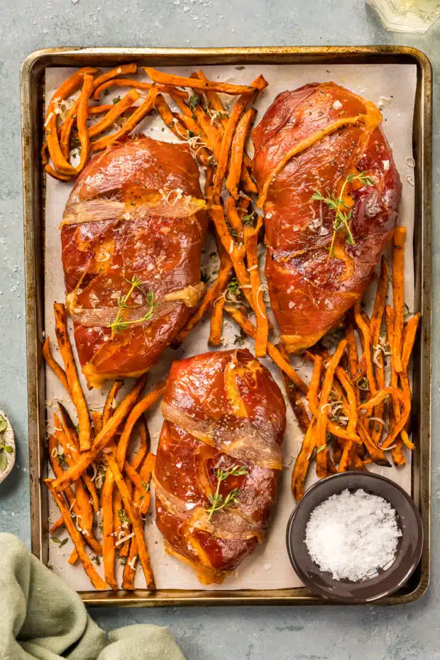 Overhead photo of three baked prosciutto wrapped chicken breasts on a sheet pan with sweet potato fries.