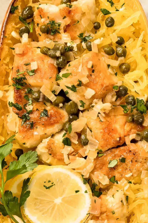 Overhead, up close shot of Healthy Chicken Piccata Spaghetti Squash Boats garnished with shredded parmesan, a slice of lemon and sprig of fresh parsley.